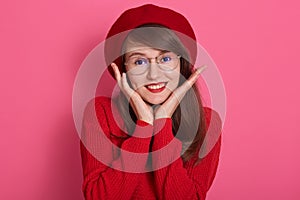 Close up portrait of smiling beautiful woman, expresses happyness, adorable girl with rounded spectacles, touching her cheeks with