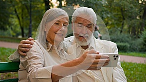 Close up portrait smiling Caucasian middle aged retired family couple talking relaxing using smartphone outdoors city