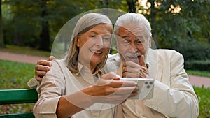 Close up portrait smiling Caucasian middle aged retired family couple talking relaxing using smartphone outdoors city