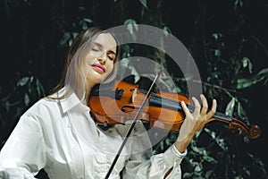Close up portrait of smiling Caucasian woman playing violin in tropical forest. Closed eyes. Music and art concept. Female with