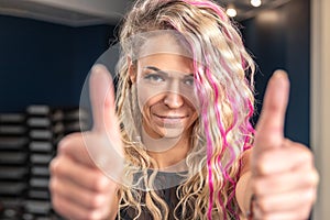 Close-up portrait of smiling fitness woman with blond and pink long hair showing thumbs up gesture. Blurred background of gym