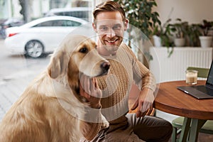 Close up portrait of smiling handsome european man with his dog in a cafe. Guy pets his golden retriever while working