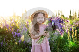 Close up portrait of a smiling little girl in a straw hat and with a large bouquet of lupins. A child girl in a field of lupines.