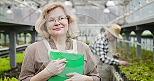 Close-up portrait of smiling senior caucasian woman in eyeglasses looking at camera as male professional agronomist