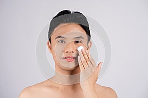 Close-up portrait of smiling shirtless young man applying facial cream, isolated on gray background. Skin care concept