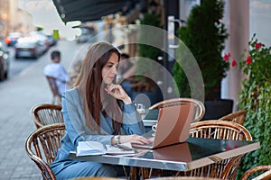 Close up portrait of smiling stylish european woman in white jacket sitting on street cafe and working on laptop.