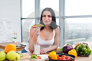 Close-up portrait of smiling woman eating diet vegetarian sandwich with vegetables for breakfast in morning, looking at