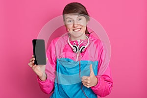 Close up portrait of smiling woman holding phone with blank screen and showing ok sign with her thu, girl has ponytail, wearing