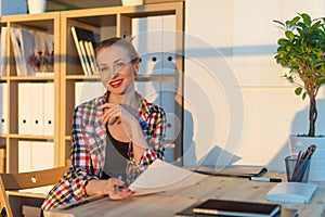 Close-up portrait of smiling woman reading paper at her workplace, looking camera on sunny day in light office room.