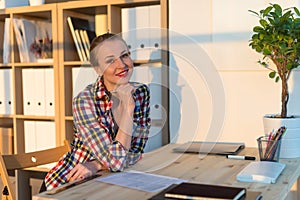 Close-up portrait of smiling woman reading paper at her workplace, looking camera on sunny day in light office room.