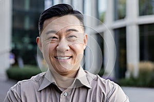 Close-up portrait of a smiling young Asian man in a brown shirt standing on a city street and looking confidently into