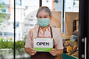 Close up portrait smiling young Asian woman in a beige shirt with a apron wearing a face mask holding an open sign board in front