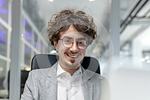 Close-up portrait of a smiling young businessman, developer man in a suit sitting in a modern office at a desk and