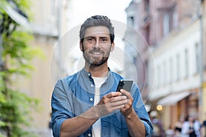 Close-up portrait of a smiling young man in a blue shirt standing on a city street, holding a mobile phone and looking