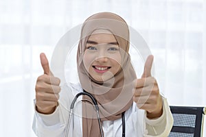 Close up portrait of smiling young muslim female medical doctor with stethoscope showing thumbs up sign in clinic