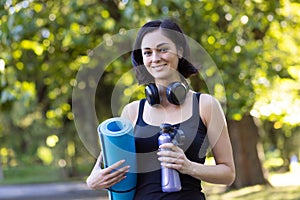 Close-up portrait of a smiling young woman standing in a park in sportswear and headphones, holding a mat and a bottle