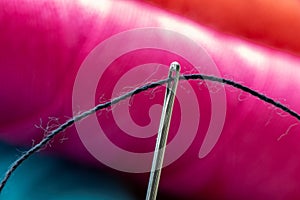 A close up portrait of some sewing thread put through the hole of a needle in front of an out of focus spool or roll of yarn. The