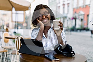 Close up portrait of stylish african american business woman, sitting at outdoor cafe with phone, tablet, headphones