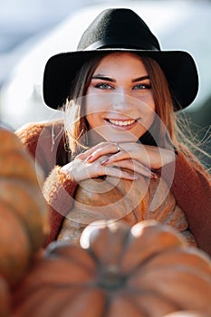 Close up portrait of stylish happy woman with smile posing among ripe orange pumpkins on farmers market in brown sweater