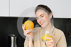 Close up portrait of stylish modern woman, drinking fresh juice from glass in kitchen, holding an orange, laughing and