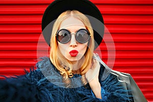 Close up portrait of stylish woman blowing red lips sending sweet air kiss stretching hand for taking selfie with shopping bags