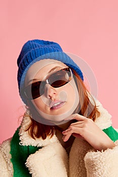 Close-up portrait of stylish young girl in sunglasses, blue hat, green scarf and fur coat posing over pink background