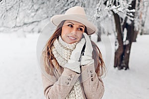 Close up portrait of stylish young woman in snowy winter park wearing fashionable white and beige warm clothes