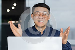 Close up portrait of successful asian man inside office, businessman using laptop at work smiling and looking at camera