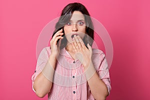 Close up portrait of surprised attractive woman with phone, tolkingwith freiendor husband, standing and covering her mouth by