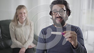 Close-up portrait of surprised Caucasian man holding pregnancy test and talking. Cheerful guy in eyeglasses happy to