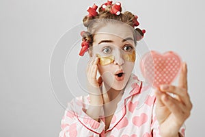 Close-up portrait of surprised or impressed young girl in hair-curlers and eye patch mask looking at heart-shaped mirror