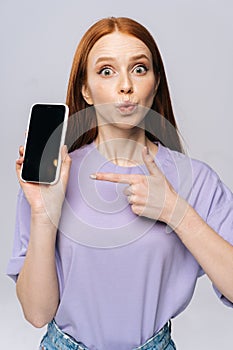 Close-up portrait of surprised young woman in casual clothes pointing with finger on blank mobile screen.