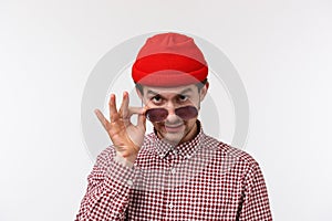 Close-up portrait of suspicious funny young guy in red beanie and checked shirt, take-off sunglasses and look from under