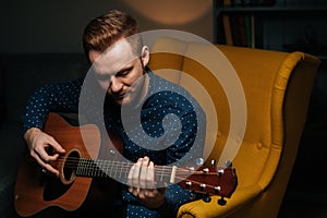 Close-up portrait of talented guitarist singer male playing acoustic guitar sitting on armchair in dark living room.