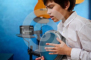 Close-up portrait teen boy with eyes closed, playing the djembe drum in a cozy home music studio, banging drums