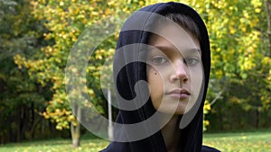 Close up portrait of teenage boy in black hoodie jacket outdoors with city park trees in background. 4K footage video