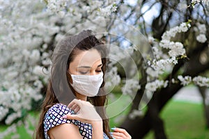 Close up portrait of tender girl in a white blouse under a blossoming cherry tree with a mask from the coronavirus