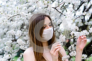 Close up portrait of tender girl in a white blouse under a blossoming cherry tree with a mask from the coronavirus