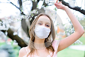 Close up portrait of tender girl in a white blouse under a blossoming cherry tree with a mask from the coronavirus