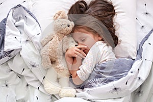 Close up portrait of toddler girl laying with her toy teddy bear in bed, posing with closed eyes, having rest after hard day, dark