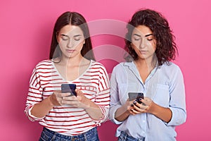 Close up portrait of two girls dress casual outfits, standing isolated over pastel pink background. Two women using mobile phones