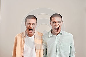 Close up portrait of two young angry twin brothers shouting and looking at camera while posing together isolated over