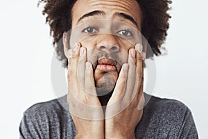 Close up portrait of upset and tired young african man grabing his face and cheeks with hands over white background
