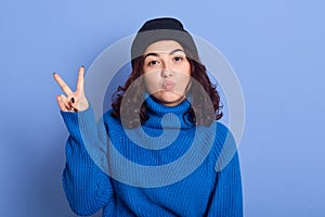 Close up portrait of winter girl wearing black cap and warm sweater, showing victory sign isolated over blue background and