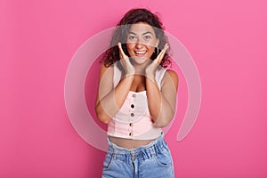 Close up portrait of woman dresses stylish pink top and jeans, holding hands on face, keeps palms near cheecks, posing with toothy