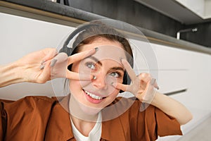 Close up portrait of woman in headphones, shows peace, v-signs near eyes, smiles and looks happy, sits on floor at home