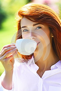 Close up portrait of a woman holding a hot coffee cup outdoors on the spring background. Red head girl drinking coffe.