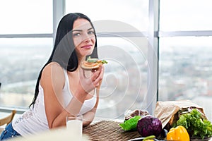 Close-up portrait of woman standing in kitchen, leaning on wooden table, having snack. Smiling girl maintains healthy