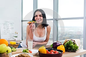 Close-up portrait of woman standing in kitchen, leaning on wooden table, having snack. Smiling girl maintains healthy