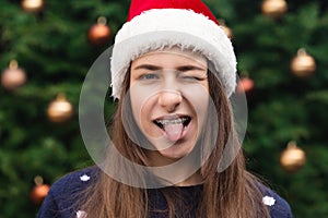 Close up Portrait of woman wearing a santa claus hat with emotion sticking tongue out. Against the background of a Christmas tree